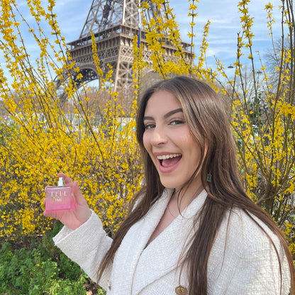 Model image of model holding ELLE In Paris by ELLE Parfums next to the Eiffel Tower. ELLE In Paris is a fruity and floral eau de parfum inspired by the elegant Parisian lifestyle. This versatile fragrance is ideal for both day and night, cruelty-free, and suitable for vegans and vegetarians. Perfect for those seeking a chic and ethical scent.