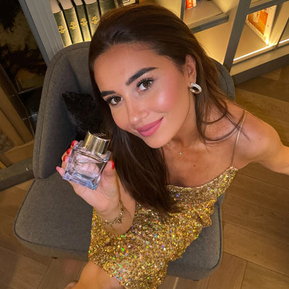 Model shot of a girl holding the ELLE Parisian Nights fragrance by ELLE Parfums in a beautiful apartment. A purple, girly bottle embodies a woody and floral scent that's a dupe for Baccarat Rouge 504 —perfect as a date night scent. This eau de perfume embodies the essence of a night out in Paris. 